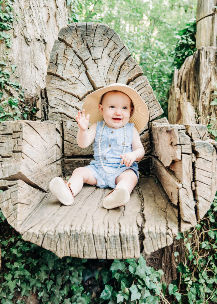 One year old girl sits in tree stump chair wearing sun hat and pearls. Farmington Utah Family Photographer.
